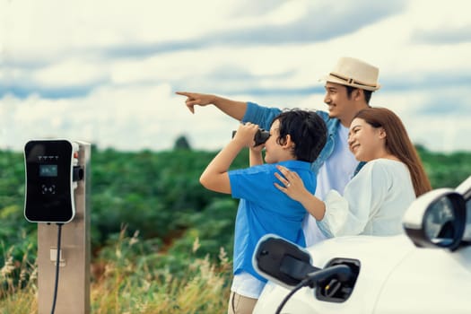 Concept of progressive happy family enjoying their time at wind farm with electric vehicle. Electric vehicle driven by clean renewable energy from wind turbine generator for charging station.