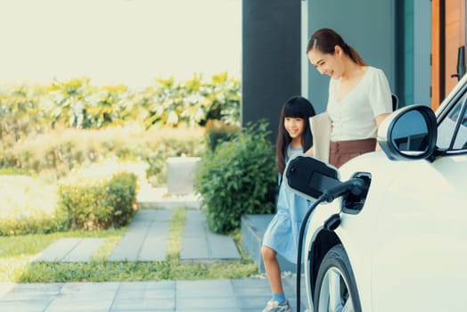 Focus electric vehicle recharging at home charging station plugged in with EV charger device with blurred background of progressive mother and daughter walking as concept for sustainability of energy.