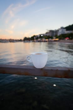 The terrace of the restaurant by the sea, a white cup of coffee stands on the pier. Against the backdrop of a beautiful resort town