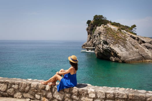 The girl in a blue dress and a straw hat alone sits near the sea on a rock, on a cliff and looks at the sea. Vacation, travel, tourism