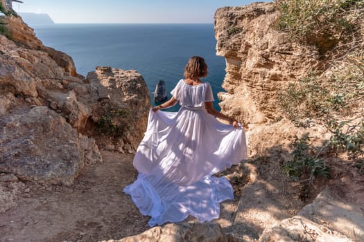 A beautiful young woman in a white light dress with long legs stands on the edge of a cliff above the sea waving a white long dress, against the background of the blue sky and the sea