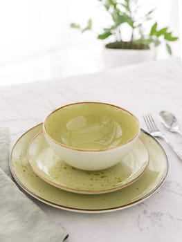 Three empty green plates with silver cutlery and tablecloth on white marble table in front of window. Minimal style table setting