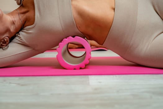 Fit athletic women in beige sportswear ride on a pink foam myofascial massage roller, massaging their muscles on the floor of a yoga mat in the gym. Active girls do a fitness workout in the gym. MFR.