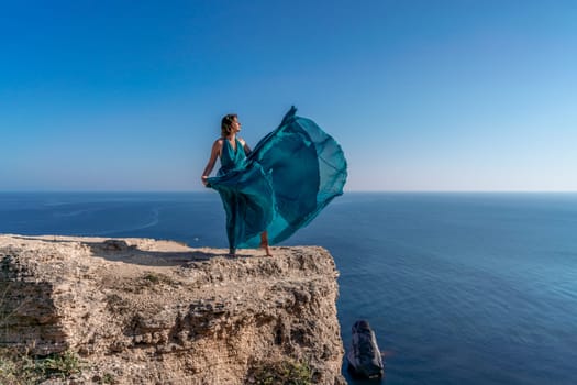 A girl with loose hair in a long mint dress descends the stairs between the yellow rocks overlooking the sea. A rock can be seen in the sea. Sunny path on the sea from the rising sun.