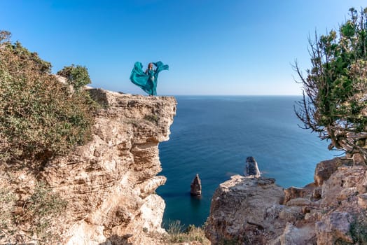 A girl with loose hair in a long mint dress descends the stairs between the yellow rocks overlooking the sea. A rock can be seen in the sea. Sunny path on the sea from the rising sun.