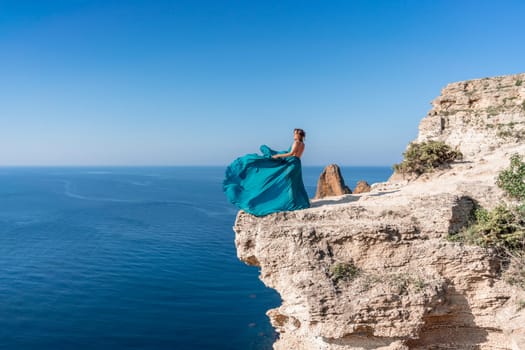 A girl with loose hair in a long mint dress descends the stairs between the yellow rocks overlooking the sea. A rock can be seen in the sea. Sunny path on the sea from the rising sun.