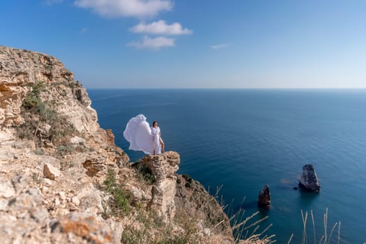 A beautiful young woman in a white light dress with long legs stands on the edge of a cliff above the sea waving a white long dress, against the background of the blue sky and the sea