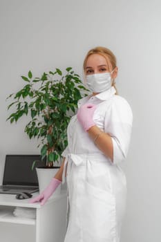 Optimistic portrait of a woman wearing a white coat. The blonde is standing next to a computer table wearing a mask and gloves. Covid19, coronavirus, the concept of healthcare and doctors