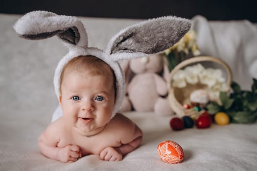 Cute little baby wearing bunny ears for Easter. Near Easter eggs. looks into the camera and smiles. High quality photo