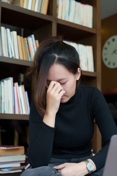 A portrait of a young Asian woman using a computer, wearing headphones and using a notebook to study online shows boredom and pain from video conferencing on a wooden desk in library.