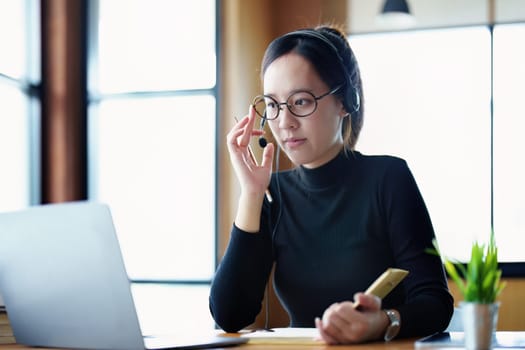 Portrait of a teenage Asian woman wearing glasses using computer laptop, headphones and using a laptop to study online via video conferencing on a wooden library table.