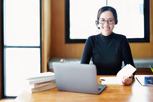 Portrait of a teenage Asian woman wearing glasses using computer laptop, headphones and using a laptop to study online via video conferencing on a wooden library table.