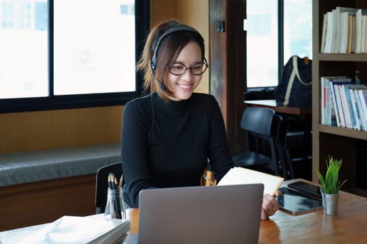 Portrait of a teenage Asian woman wearing glasses using computer laptop, headphones and using a laptop to study online via video conferencing on a wooden library table.