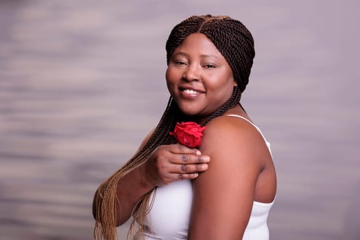 Attractive african american model posing with red rose bud. Carefree natural beautiful curvy woman with positive facial expression holding romantic symbol flower in hand portrait