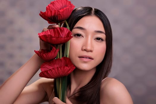 Asian young woman leaning red poppies to face portrait. Attractive lady holding romantic love symbol in hands, looking at camera and posing with beautiful bright flowers bouquet