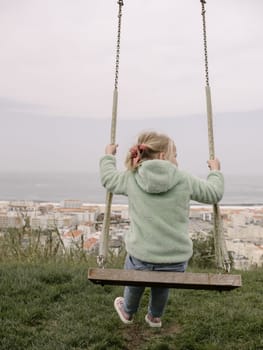 A 5-year-old girl is swinging on simple wooden swings on top of a hill, viewed from the back. In the distance, a city and ocean are visible