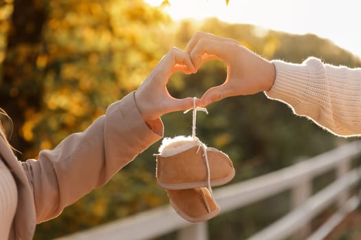 Man and pregnant wife are doing heart gesture with hands and holding warm baby shoes on nature autumn background. Future parents waiting for the baby. The concept of Mother's Day and Women's Day