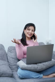 Portrait of a beautiful Asian teenage girl using her phone and computer sitting on the sofa at home.