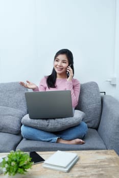 Portrait of a beautiful Asian teenage girl using her phone and computer sitting on the sofa at home.