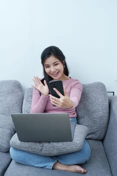 Portrait of a beautiful Asian teenage girl using her phone and computer for video conferencing sitting on the sofa at home.