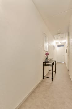 a long hallway with white walls and beige carpet on the floor, there is a small black table in the corner