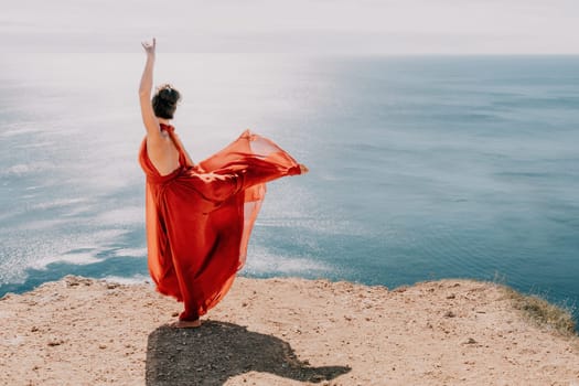 Side view a Young beautiful sensual woman in a red long dress posing on a rock high above the sea during sunrise. Girl on the nature on blue sky background. Fashion photo.