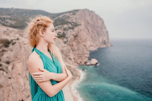 Side view a Young beautiful sensual woman in a mint long dress posing on a volcanic rock high above the sea during sunset. Girl on the nature on overcast sky background. Fashion photo