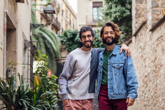 gay male couple embracing looking at camera smiling happy in a nice street with plants, concept of leisure and love between people of the same sex