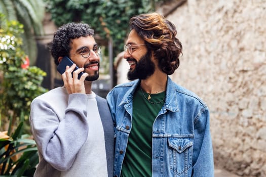 gay male couple smiling happily while one of them is talking on the phone in a nice street with plants, concept of communication and love between people of the same sex