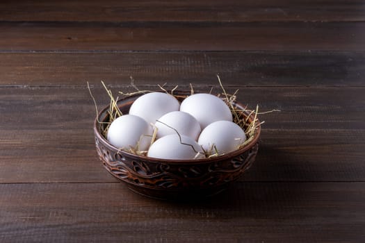 White raw chicken eggs lie on hay in a burnt clay bowl on the surface of their natural dark wood. Healthy food. Dark key, rustic.