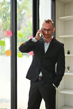 Portrait of caucasian man corporate CEO in black suit standing in modern office and talking on mobile phone.