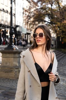 charming brunette posing on the street in spring, wearing a stylish beige coat, a top with razors and sunglasses