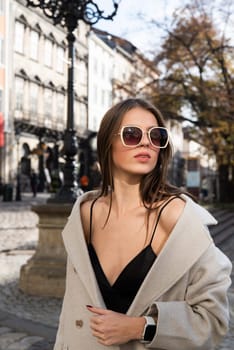 charming brunette posing on the street in spring, wearing a stylish beige coat, a top with razors and sunglasses