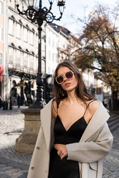 charming brunette posing on the street in spring, wearing a stylish beige coat, a top with razors and sunglasses