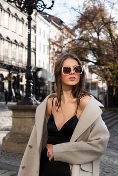 charming brunette posing on the street in spring, wearing a stylish beige coat, a top with razors and sunglasses