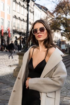 charming brunette posing on the street in spring, wearing a stylish beige coat, a top with razors and sunglasses