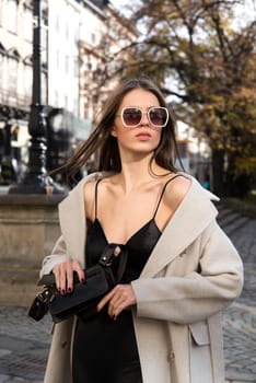 charming brunette posing on the street in spring, wearing a stylish beige coat, a top with razors and sunglasses