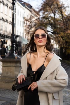 charming brunette posing on the street in spring, wearing a stylish beige coat, a top with razors and sunglasses