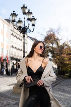 charming brunette posing on the street in spring, wearing a stylish beige coat, a top with razors and sunglasses