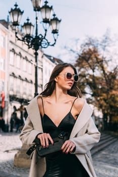 charming brunette posing on the street in spring, wearing a stylish beige coat, a top with razors and sunglasses