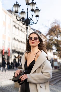 charming brunette posing on the street in spring, wearing a stylish beige coat, a top with razors and sunglasses