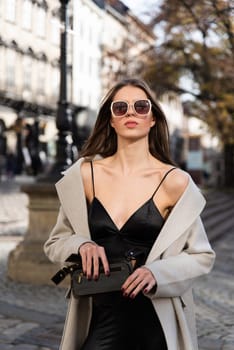 charming brunette posing on the street in spring, wearing a stylish beige coat, a top with razors and sunglasses