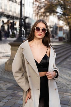 charming brunette posing on the street in spring, wearing a stylish beige coat, a top with razors and sunglasses