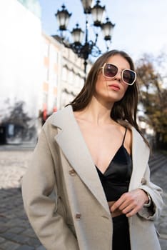 charming brunette posing on the street in spring, wearing a stylish beige coat, a top with razors and sunglasses