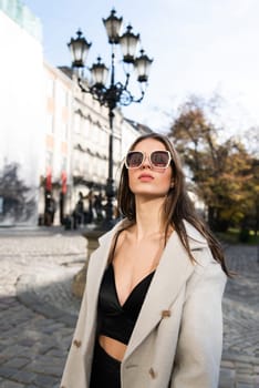 charming brunette posing on the street in spring, wearing a stylish beige coat, a top with razors and sunglasses