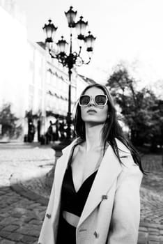 charming brunette posing on the street in spring, wearing a stylish beige coat, a top with razors and sunglasses