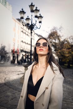 charming brunette posing on the street in spring, wearing a stylish beige coat, a top with razors and sunglasses