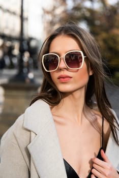 charming brunette posing on the street in spring, wearing a stylish beige coat, a top with razors and sunglasses