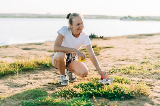 Young female volunteer satisfied with picking up trash, a plastic bottles and coffee cups, clean up beach with a sea. Woman collecting garbage. Environmental ecology pollution concept. Earth Day.