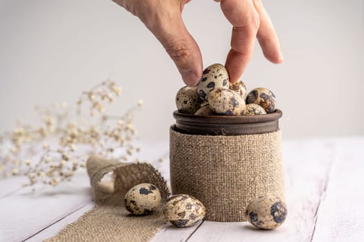 The hand takes a quail egg. Fresh quail eggs in a clay mug on the table. Bright spring festive background and a branch of gypsophila. Close-up. Healthy eating. Easter